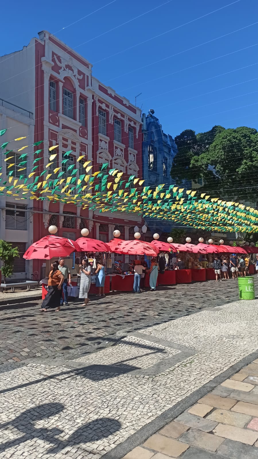 Barracas com guarda-sóis vermelhos da Feira do Bom Jesus, no Recife Antigo, sob bandeirinhas verdes e amarelas e casarões coloridos ao fundo